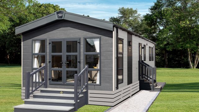 Glade with green lawn illuminated by sunlight. Wavy landscape in the background with tall trees.